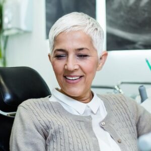 An older woman with short white hair smiles while seated at the dentist